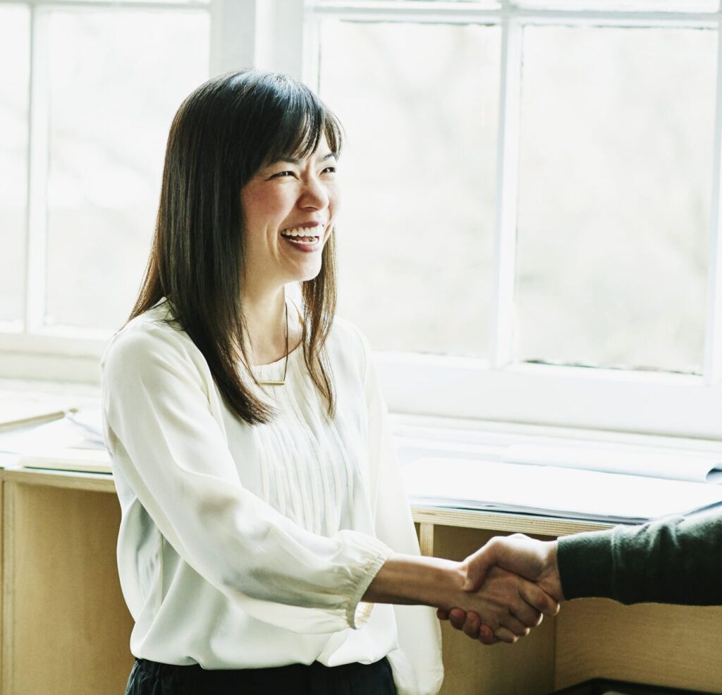 A woman in a white blouse is smiling and shaking hands with another person. They are standing in a bright room with large windows in the background.